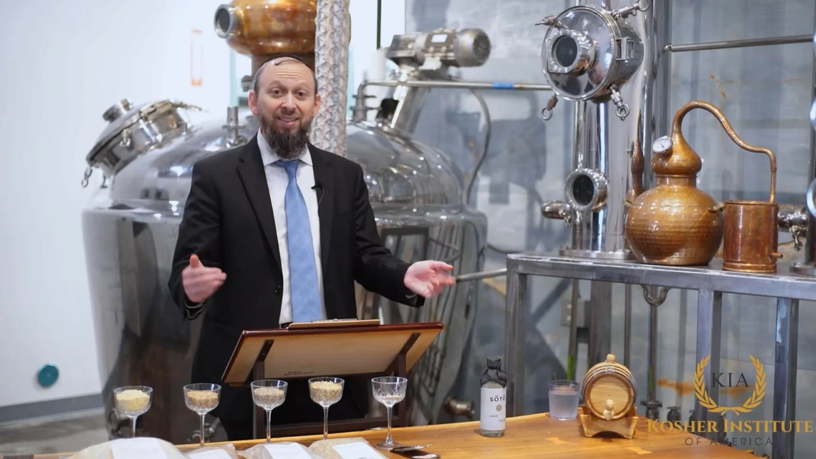 Rabbi Akiva Niehaus teaches the Kosher Liquor Masterclass at a lectern on a distillery floor, tasting glasses of grain lined up in front of him.